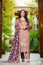 "An elegant model is wearing an embroidered mauve three-piece suit. The dress has delicate embroidery on the neckline and bottom, and is paired with a blue and mauve printed dupatta. A traditional structure and lush green plants are visible in the background."